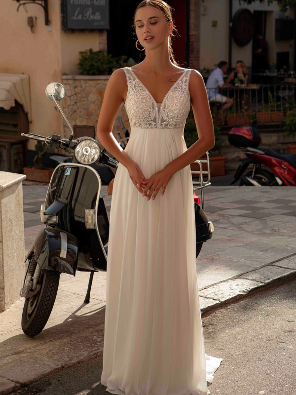 Mariée debout devant un scooter vintage noir, vêtue d'une robe fluide avec un corsage en dentelle et un décolleté en V, dans un cadre de village ensoleillé