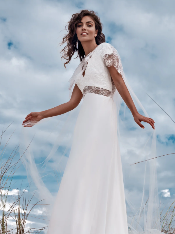 Mariée élégante dans une robe fluide blanche avec des motifs en dentelle, debout sur une dune de sable avec des herbes sauvages et un ciel nuageux en arrière-plan