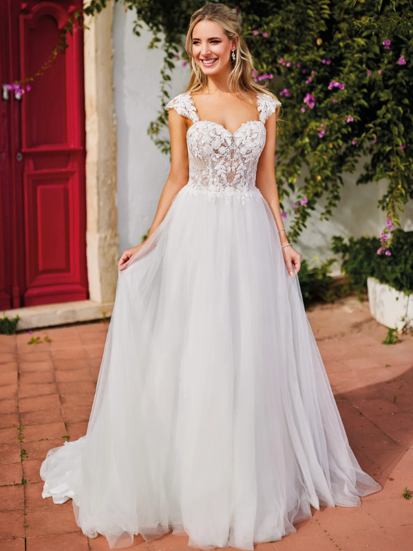 Mariée radieuse dans une robe de mariée au corsage floral en dentelle, posant devant une porte rouge entourée de végétation luxuriante et de fleurs violettes