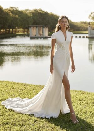 Une mariée élégante portant une robe de mariée blanche minimaliste avec des manches courtes et une fente sur la jupe, posant près d’un lac entouré de verdure.