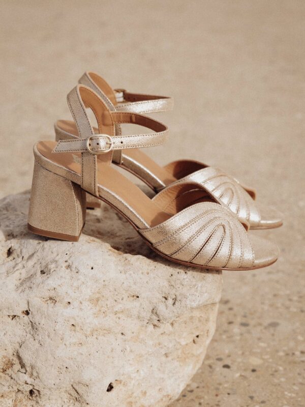 Sandales dorées à talon épais posées sur une pierre blanche, mettant en valeur leur design raffiné et éclatant.