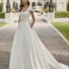 Une mariée élégante portant une robe de mariée princesse avec un col carré structuré et une jupe ample, posant dans une allée bordée d’un château majestueux.
