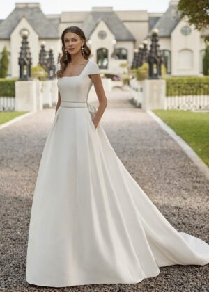 Une mariée élégante portant une robe de mariée princesse avec un col carré structuré et une jupe ample, posant dans une allée bordée d’un château majestueux.