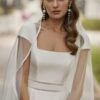 Gros plan sur une mariée portant une robe de mariée à col carré avec une cape en voile délicate et des boucles d’oreilles pendantes.