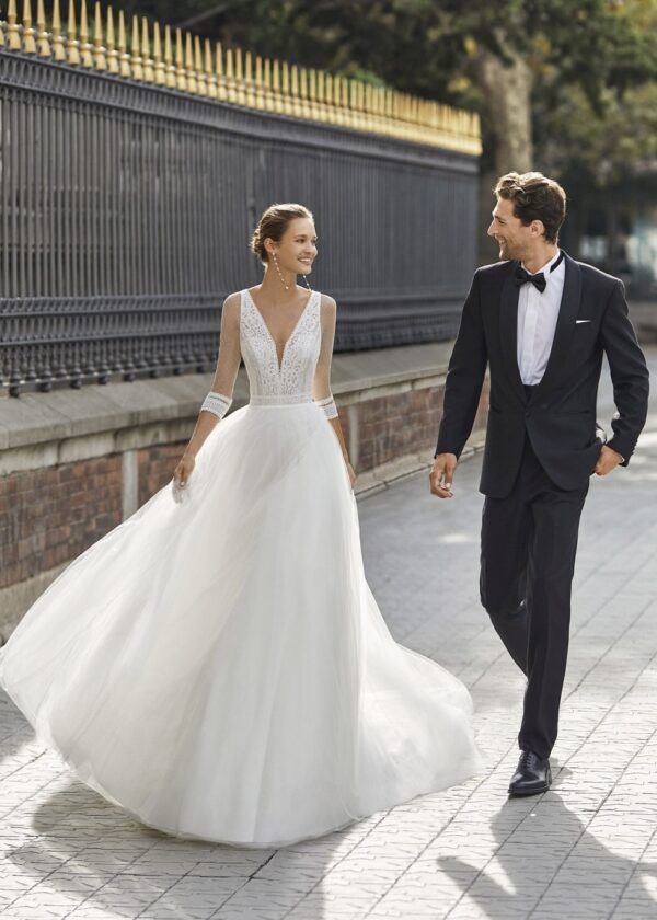 Une mariée en robe blanche à jupe volumineuse marchant joyeusement aux côtés de son partenaire en costume noir élégant devant une grille dorée.