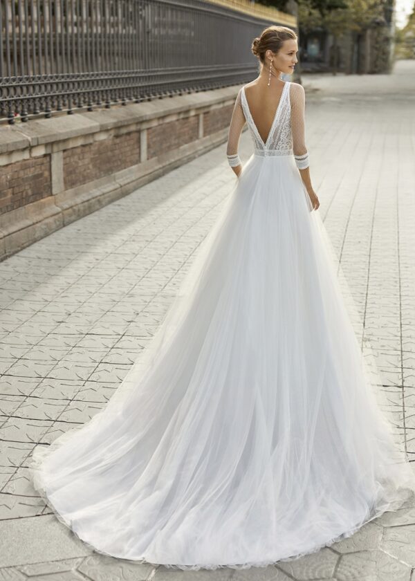 Une vue arrière d'une robe de mariée avec une longue traîne en tulle, un dos nu en V orné de dentelle et des manches longues élégantes, posant sur une rue pavée bordée d'une grille historique.