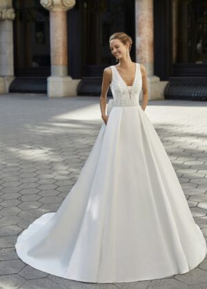 Une femme élégante portant une robe de mariée blanche avec une jupe volumineuse, un décolleté en V en dentelle et un corsage ajusté, debout sur une place pavée.