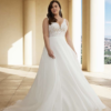 Une mariée élégante dans une robe à décolleté en V avec un corsage en dentelle et une jupe fluide, posant sur une terrasse ensoleillée entourée de colonnes, avec une vue panoramique en arrière-plan