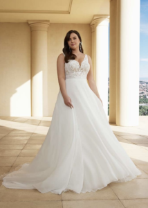 Une mariée élégante dans une robe à décolleté en V avec un corsage en dentelle et une jupe fluide, posant sur une terrasse ensoleillée entourée de colonnes, avec une vue panoramique en arrière-plan