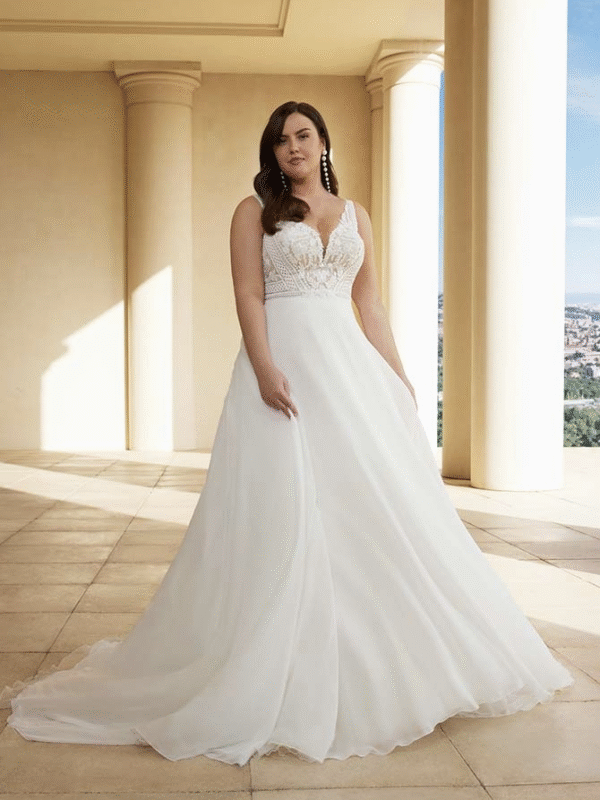 Une mariée élégante dans une robe à décolleté en V avec un corsage en dentelle et une jupe fluide, posant sur une terrasse ensoleillée entourée de colonnes, avec une vue panoramique en arrière-plan