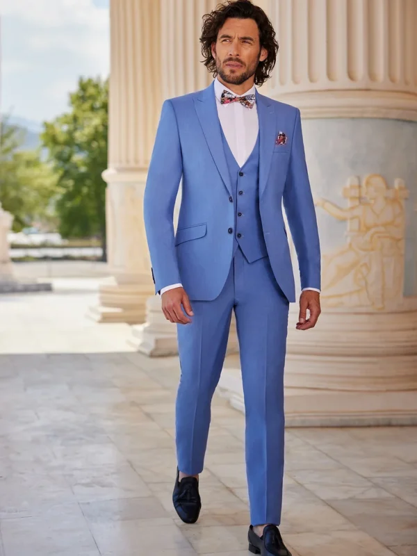 Homme habillé d’un costume bleu ciel élégant, assorti d’un gilet et d’un nœud papillon coloré, marchant dans un cadre classique avec des colonnes sculptées.