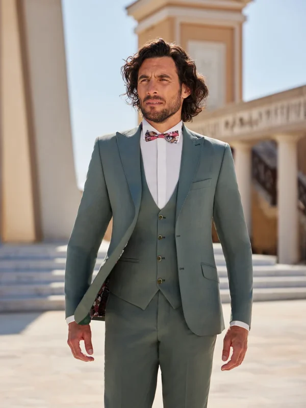 Homme portant un costume vert olive élégant avec un gilet assorti et un nœud papillon à motifs colorés, se tenant devant une architecture classique baignée de lumière