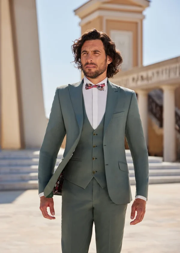 Homme portant un costume vert olive élégant avec un gilet assorti et un nœud papillon à motifs colorés, se tenant devant une architecture classique baignée de lumière