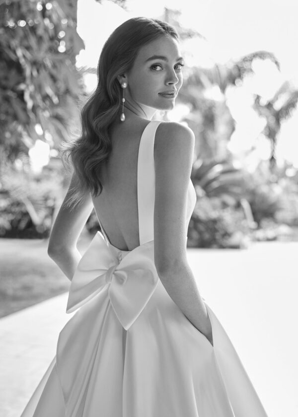 Portrait d’une mariée vue de dos portant une robe de mariée satinée avec un grand nœud et un décolleté profond dans un cadre tropical.
