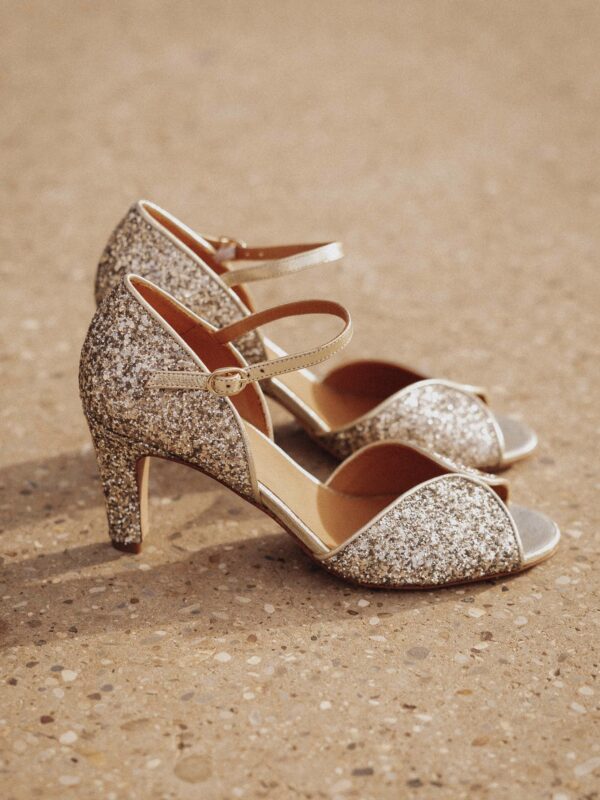 Une paire d’escarpins dorés à paillettes avec talon moyen et bride à la cheville, posée sur un sol texturé.