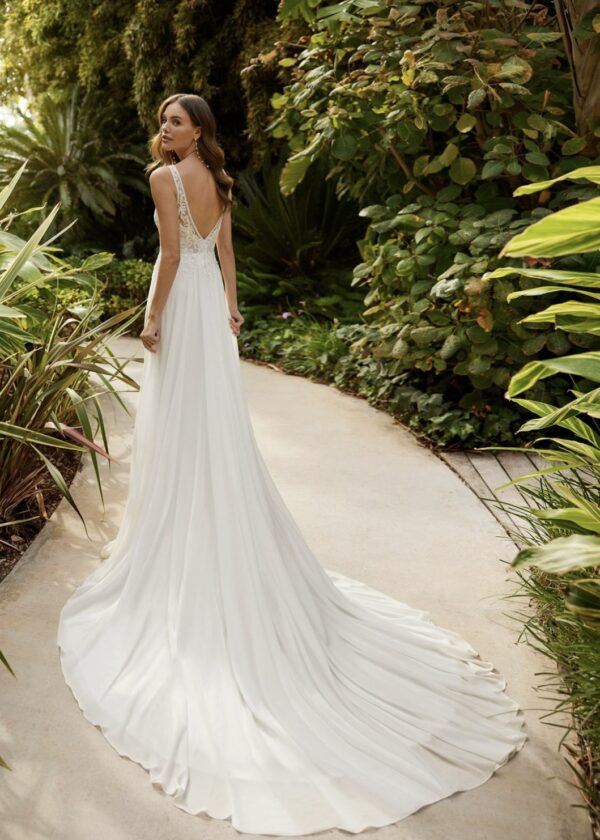 Mariée de dos portant une robe de mariée fluide avec un dos en V plongeant et une longue traîne, dans un cadre naturel luxuriant