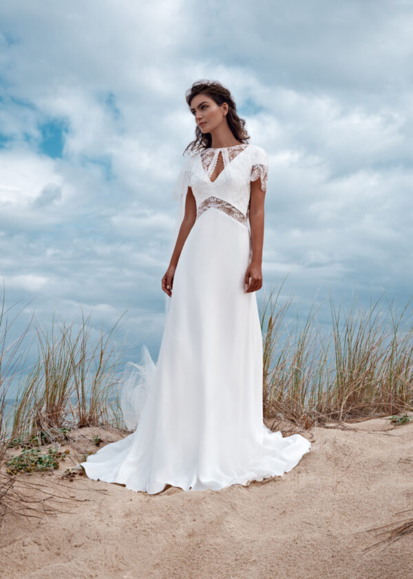 Mariée élégante vêtue d'une robe fluide avec des détails en dentelle délicate, posant dans un paysage naturel avec un ciel nuageux et des herbes hautes en arrière-plan.