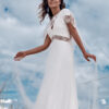 Mariée élégante dans une robe fluide blanche avec des motifs en dentelle, debout sur une dune de sable avec des herbes sauvages et un ciel nuageux en arrière-plan