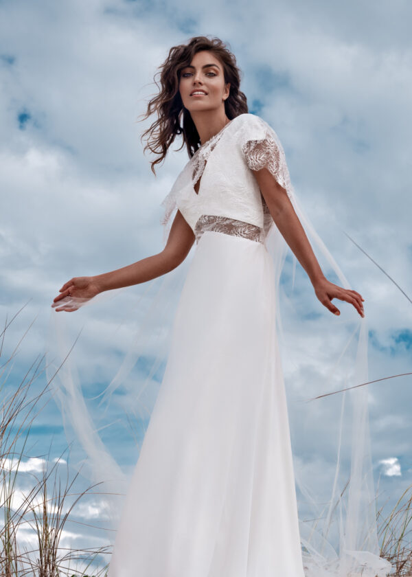 Mariée élégante dans une robe fluide blanche avec des motifs en dentelle, debout sur une dune de sable avec des herbes sauvages et un ciel nuageux en arrière-plan