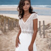 Jeune femme portant une robe de mariée en dentelle délicate avec des manches courtes et un décolleté en V, posant sur une plage au bord de l'océan