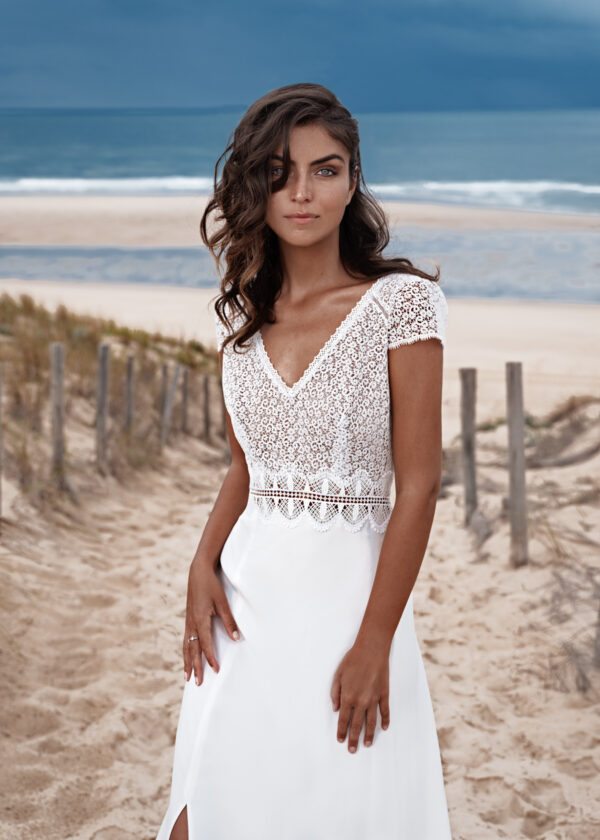 Jeune femme portant une robe de mariée en dentelle délicate avec des manches courtes et un décolleté en V, posant sur une plage au bord de l'océan