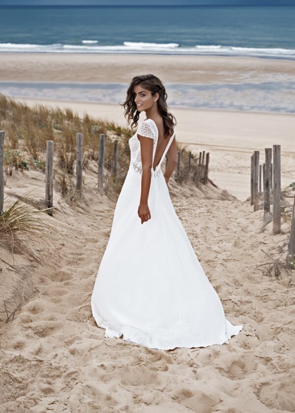 Mariée souriante en robe blanche avec un dos ouvert élégant, marchant sur un chemin de sable avec une vue sur la plage et l'océan en arrière-plan