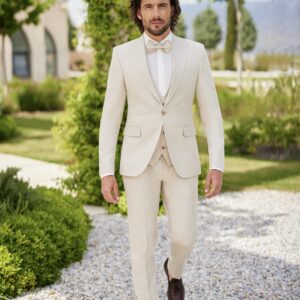 Homme portant un costume beige clair élégant avec un nœud papillon assorti, marchant dans un jardin verdoyant avec des éléments architecturaux en arrière-plan.