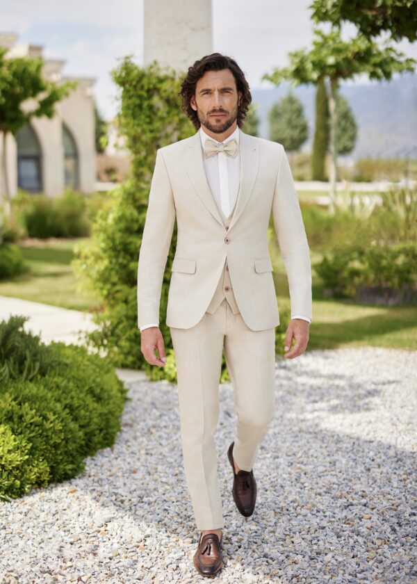 Homme portant un costume beige clair élégant avec un nœud papillon assorti, marchant dans un jardin verdoyant avec des éléments architecturaux en arrière-plan.