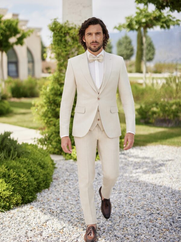 Homme portant un costume beige clair élégant avec un nœud papillon assorti, marchant dans un jardin verdoyant avec des éléments architecturaux en arrière-plan.