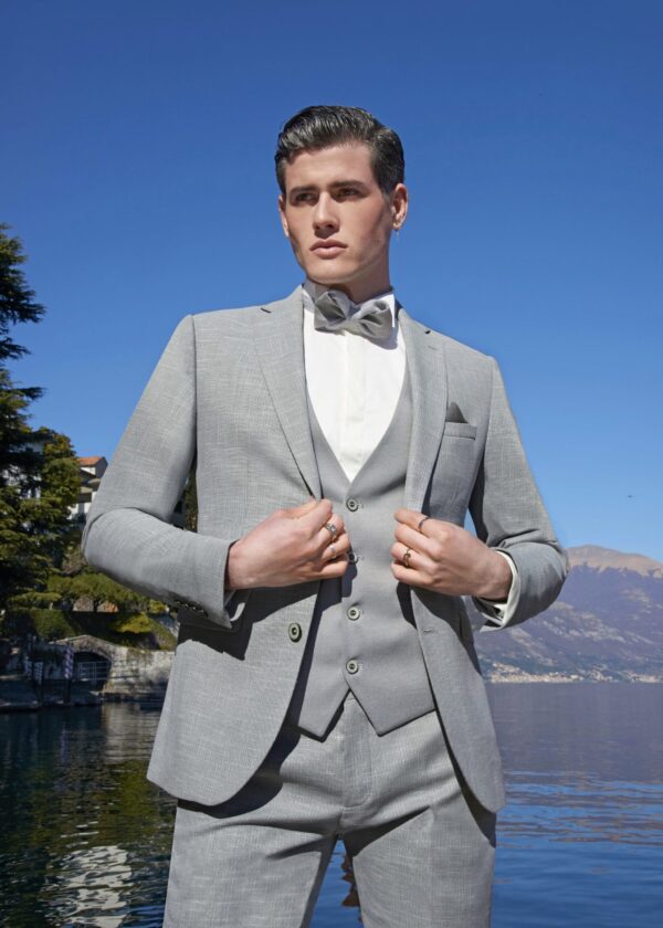 Homme vêtu d’un costume gris clair avec un gilet assorti et un nœud papillon, posant devant un lac scintillant et un ciel bleu dégagé.