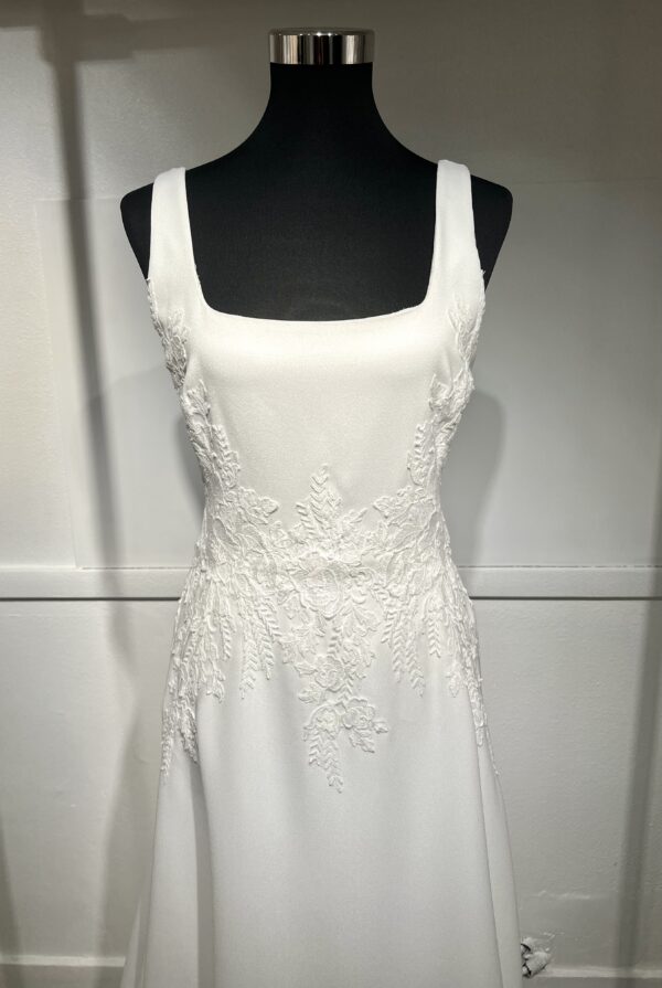 Cette robe de mariée allie minimalisme et élégance avec son corsage ajusté à col carré et sa jupe fluide prolongée d’une traîne légère. Son design épuré s’harmonise parfaitement avec un cadre méditerranéen raffiné, idéal pour une mariée au style intemporel.