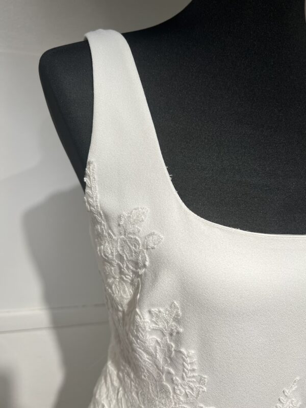 Cette robe de mariée allie minimalisme et élégance avec son corsage ajusté à col carré et sa jupe fluide prolongée d’une traîne légère. Son design épuré s’harmonise parfaitement avec un cadre méditerranéen raffiné, idéal pour une mariée au style intemporel.