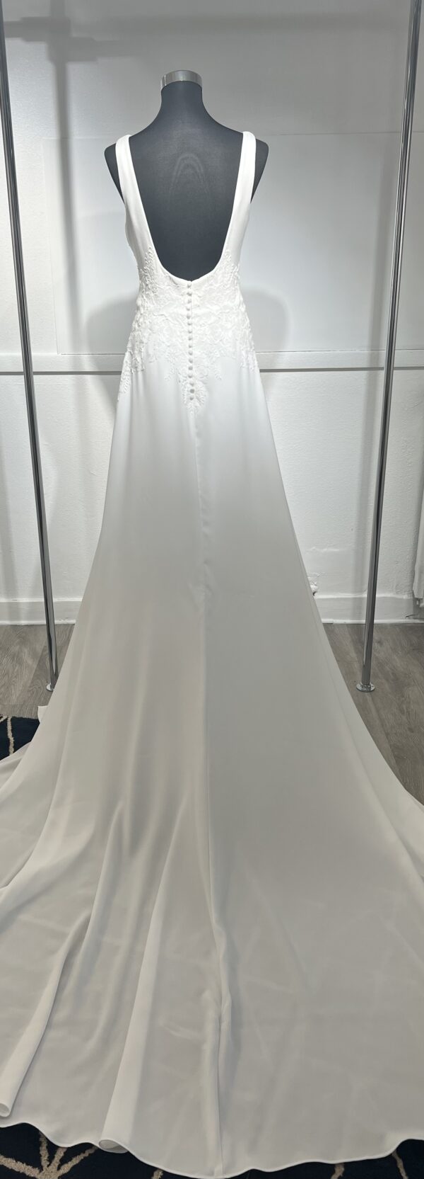 Cette robe de mariée allie minimalisme et élégance avec son corsage ajusté à col carré et sa jupe fluide prolongée d’une traîne légère. Son design épuré s’harmonise parfaitement avec un cadre méditerranéen raffiné, idéal pour une mariée au style intemporel.