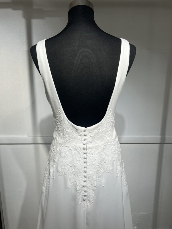Cette robe de mariée allie minimalisme et élégance avec son corsage ajusté à col carré et sa jupe fluide prolongée d’une traîne légère. Son design épuré s’harmonise parfaitement avec un cadre méditerranéen raffiné, idéal pour une mariée au style intemporel.