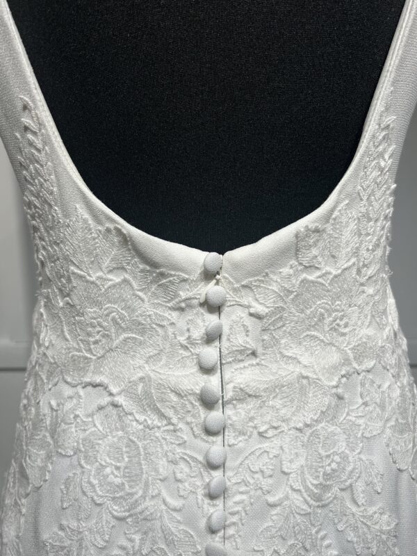 Cette robe de mariée allie minimalisme et élégance avec son corsage ajusté à col carré et sa jupe fluide prolongée d’une traîne légère. Son design épuré s’harmonise parfaitement avec un cadre méditerranéen raffiné, idéal pour une mariée au style intemporel.