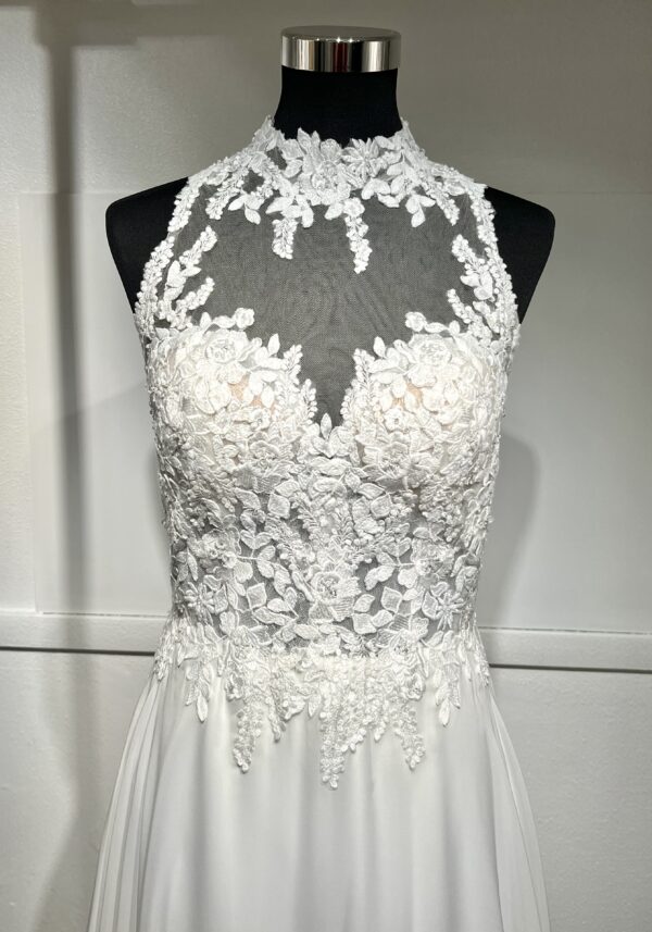 Cette robe de mariée allie raffinement et modernité avec son corsage en dentelle florale et son col haut élégant. Sa jupe fluide crée une silhouette aérienne, sublimée par un décor contemporain. Parfaite pour une mariée en quête de romantisme et de minimalisme.