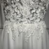 Cette robe de mariée allie raffinement et modernité avec son corsage en dentelle florale et son col haut élégant. Sa jupe fluide crée une silhouette aérienne, sublimée par un décor contemporain. Parfaite pour une mariée en quête de romantisme et de minimalisme.