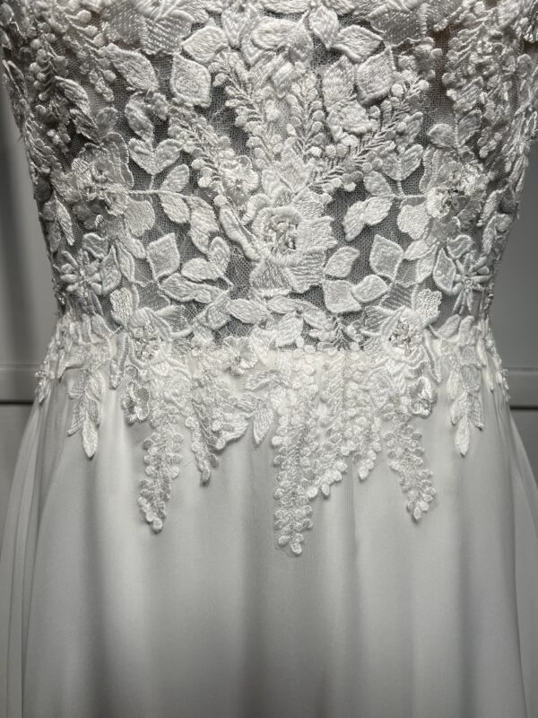 Cette robe de mariée allie raffinement et modernité avec son corsage en dentelle florale et son col haut élégant. Sa jupe fluide crée une silhouette aérienne, sublimée par un décor contemporain. Parfaite pour une mariée en quête de romantisme et de minimalisme.