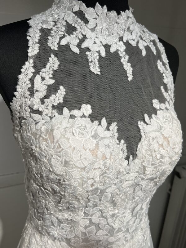 Cette robe de mariée allie raffinement et modernité avec son corsage en dentelle florale et son col haut élégant. Sa jupe fluide crée une silhouette aérienne, sublimée par un décor contemporain. Parfaite pour une mariée en quête de romantisme et de minimalisme.