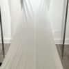 Une robe de mariée blanche minimaliste, avec un haut fluide sans manches et une jupe droite. Elle se distingue par sa simplicité élégante, mise en valeur dans un couloir moderne en pierre.