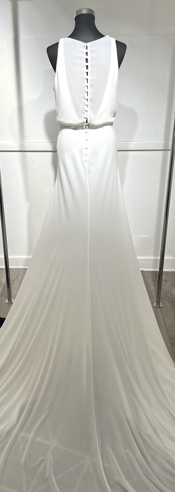 Une robe de mariée blanche minimaliste, avec un haut fluide sans manches et une jupe droite. Elle se distingue par sa simplicité élégante, mise en valeur dans un couloir moderne en pierre.
