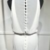 Une robe de mariée blanche minimaliste, avec un haut fluide sans manches et une jupe droite. Elle se distingue par sa simplicité élégante, mise en valeur dans un couloir moderne en pierre.