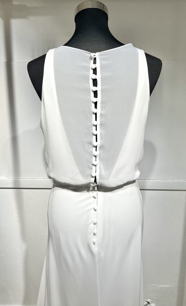 Une robe de mariée blanche minimaliste, avec un haut fluide sans manches et une jupe droite. Elle se distingue par sa simplicité élégante, mise en valeur dans un couloir moderne en pierre.
