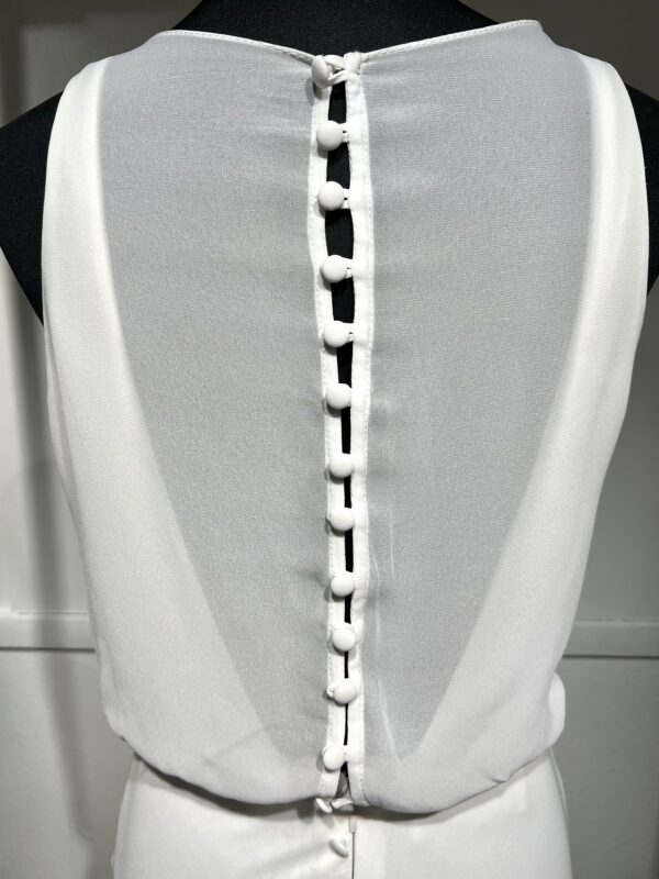 Une robe de mariée blanche minimaliste, avec un haut fluide sans manches et une jupe droite. Elle se distingue par sa simplicité élégante, mise en valeur dans un couloir moderne en pierre.