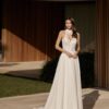 Une mariée portant une robe blanche avec un corsage en dentelle florale et un col haut, posant élégamment devant une villa moderne en bois.