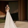 Vue arrière d'une robe de mariée fluide avec un dos en dentelle transparente et une longue traîne élégante, posant dans un cadre moderne aux finitions en bois.