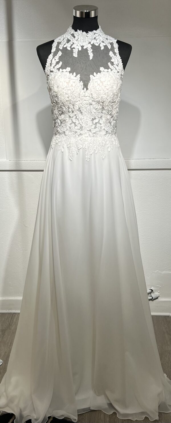 Cette robe de mariée allie raffinement et modernité avec son corsage en dentelle florale et son col haut élégant. Sa jupe fluide crée une silhouette aérienne, sublimée par un décor contemporain. Parfaite pour une mariée en quête de romantisme et de minimalisme.