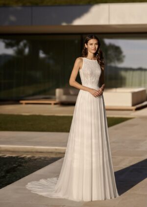 Une mariée portant une robe blanche minimaliste avec un corsage en dentelle et une jupe fluide plissée, posant dans un cadre moderne avec une villa élégante en arrière-plan.