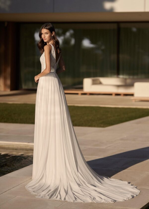 Une vue arrière d'une robe de mariée fluide avec un dos échancré et une longue traîne plissée, posant dans un cadre extérieur élégant et moderne.