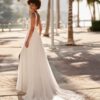 Une mariée se tient de profil, vêtue d’une robe de mariée en dentelle avec un dos échancré et une jupe fluide traînante, dans une promenade bordée de palmiers et baignée de lumière naturelle